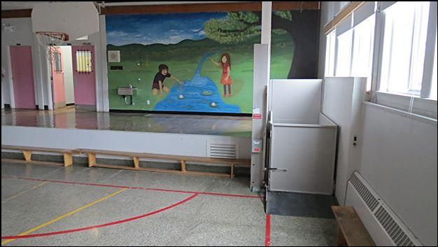 Cafétéria scolaire vide avec de longs bancs, une fresque murale et des fenêtres sur le côté droit.