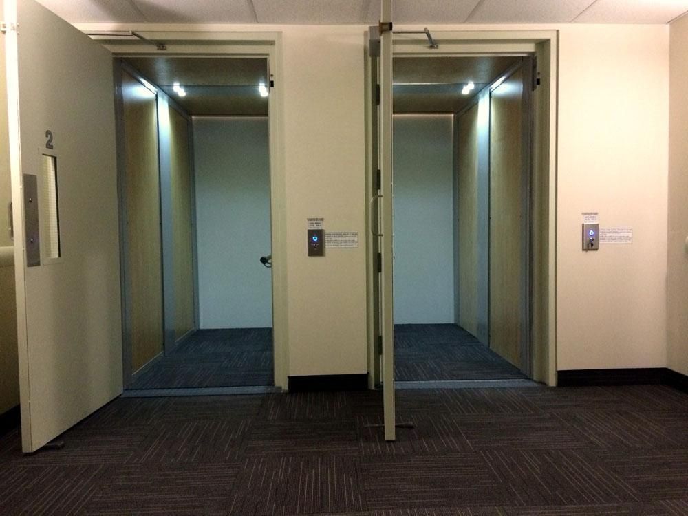 Deux ascenseurs ouverts dans un couloir beige au sol recouvert de moquette foncée.
