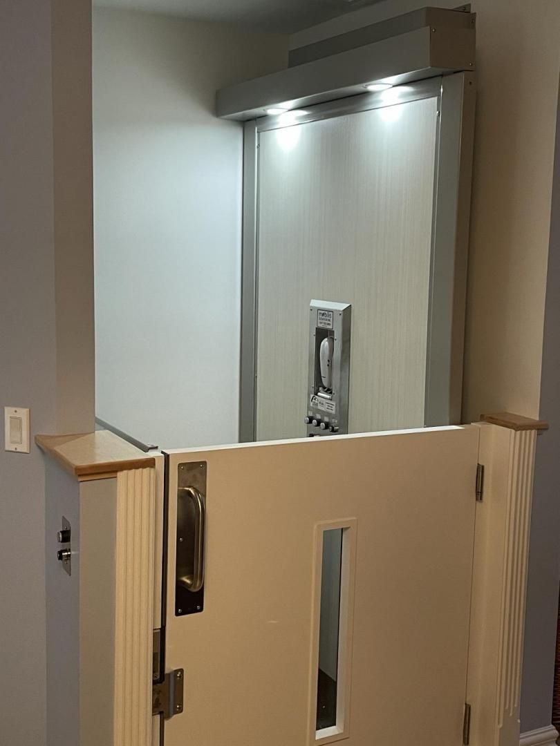 Porte beige entrouverte avec une fenêtre étroite, laissant entrevoir l'intérieur d'un ascenseur au-delà d'une barrière de couloir.