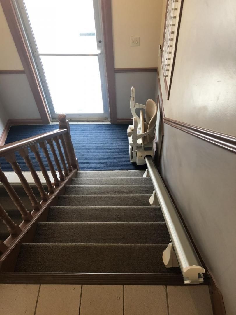 Cage d'escalier avec marches recouvertes de moquette, rampe en bois, main courante murale et porte en bas laissant entrer la lumière du jour.
