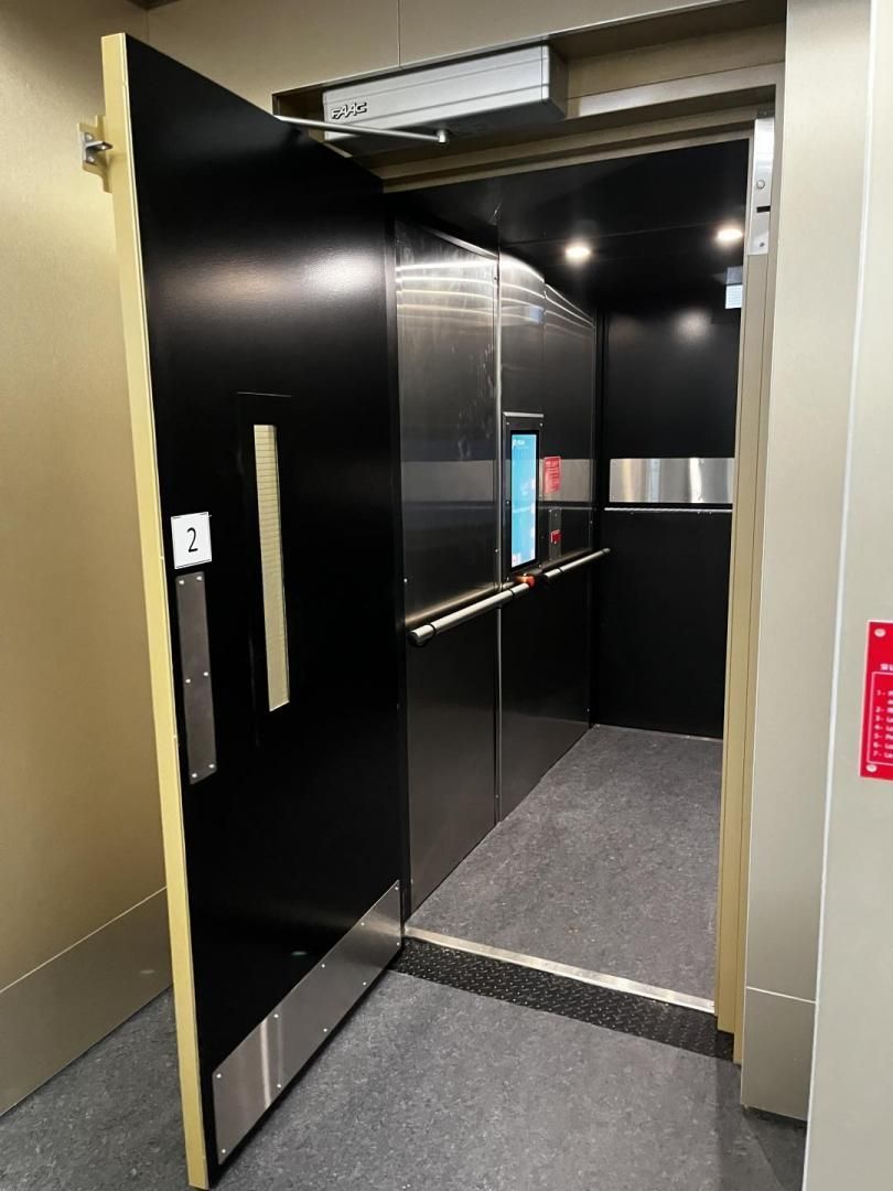 Ascenseur ouvert avec portes noires, rampe argentée et moquette grise dans un couloir.