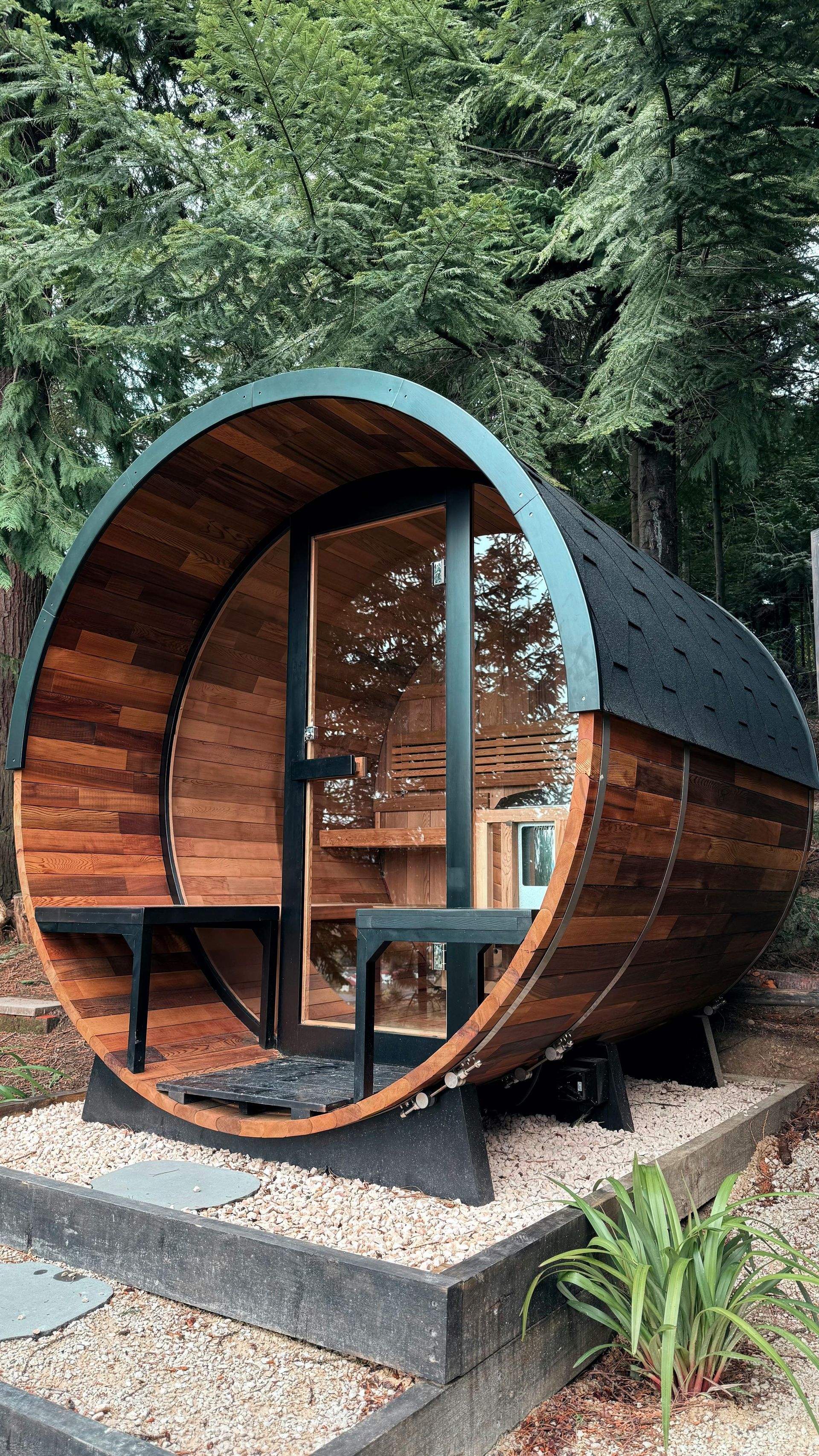 Wooden barrel-shaped structure with a glass door, benches inside. Set on gravel with greenery and trees in the background.