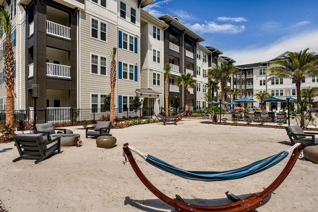 A sandy courtyard with hammocks and lounge seating, surrounded by modern apartment buildings and palm trees under a clear blue sky at Isla Antigua, offers apartments for rent in St. Augustine, FL.