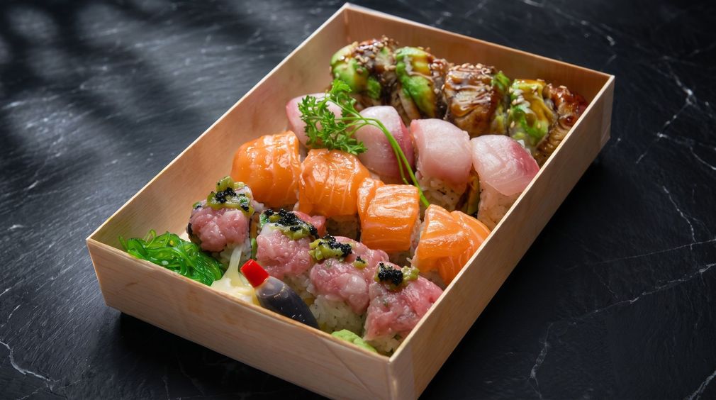 Assorted sushi rolls in a takeout tray, topped with sauces and garnish