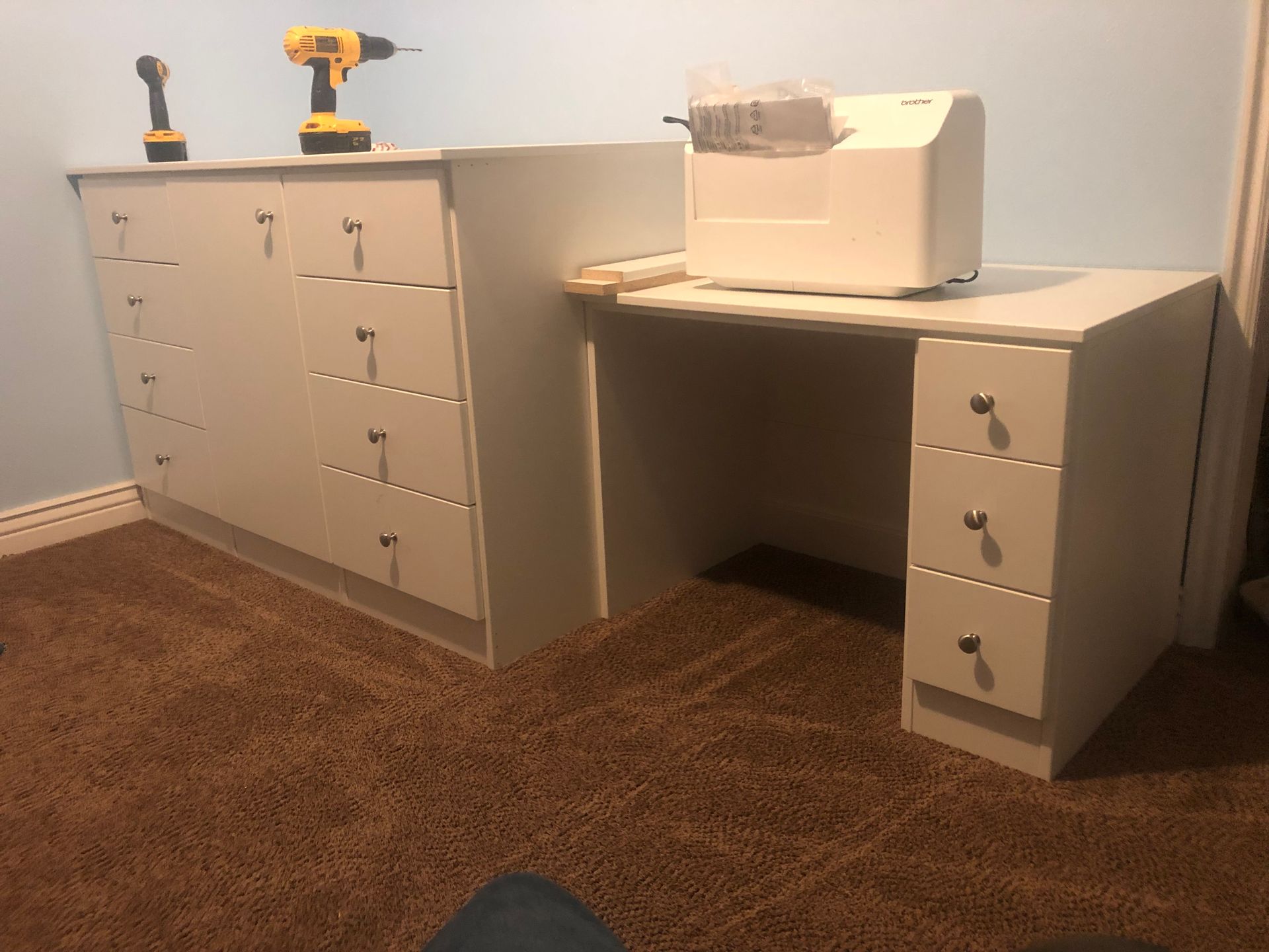A white desk with drawers and a sewing machine.  A wall unit with drawers and power tools. On a brown carpet.