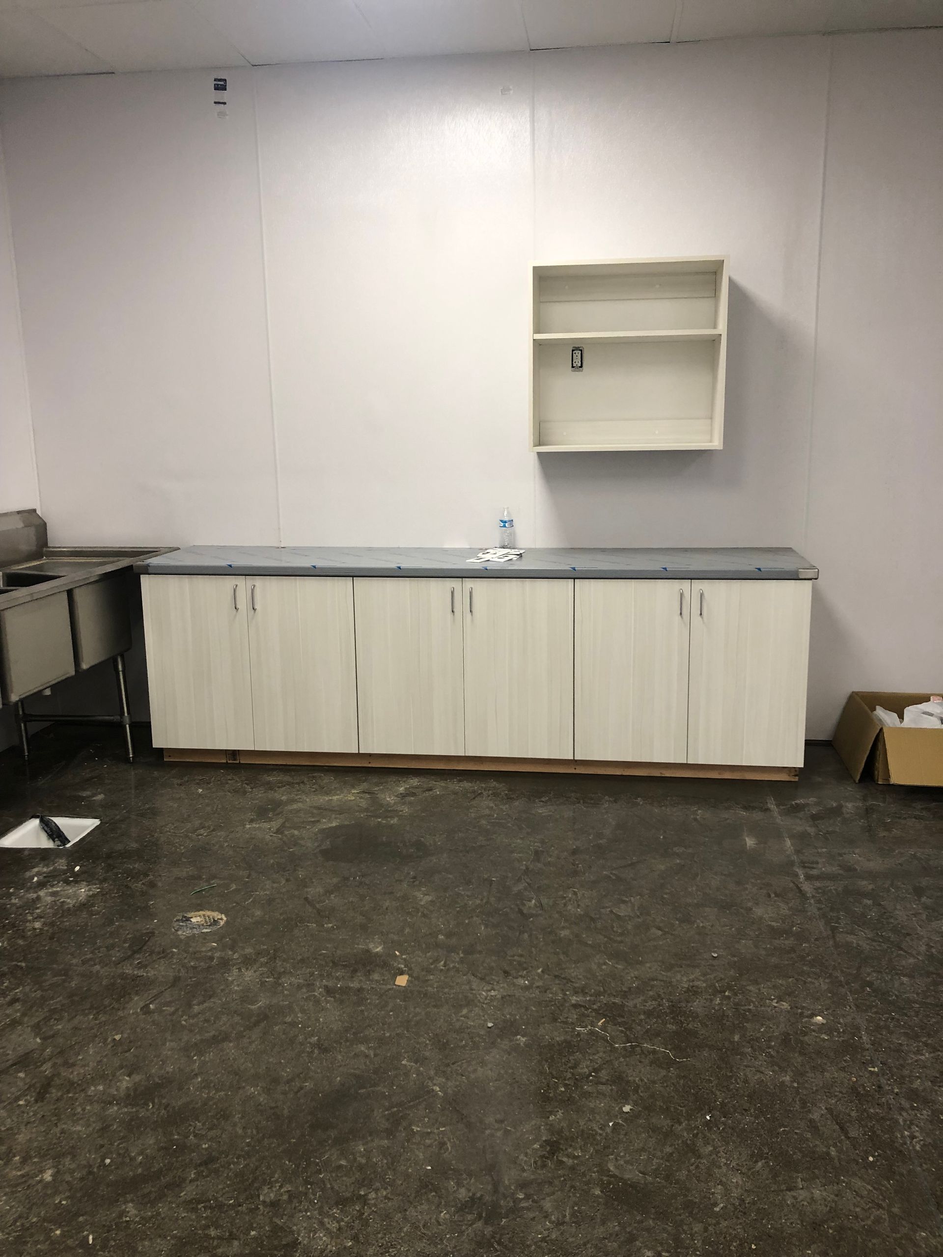 Empty room with white cabinets, countertop, and upper shelf. Dirty floor, white wall.