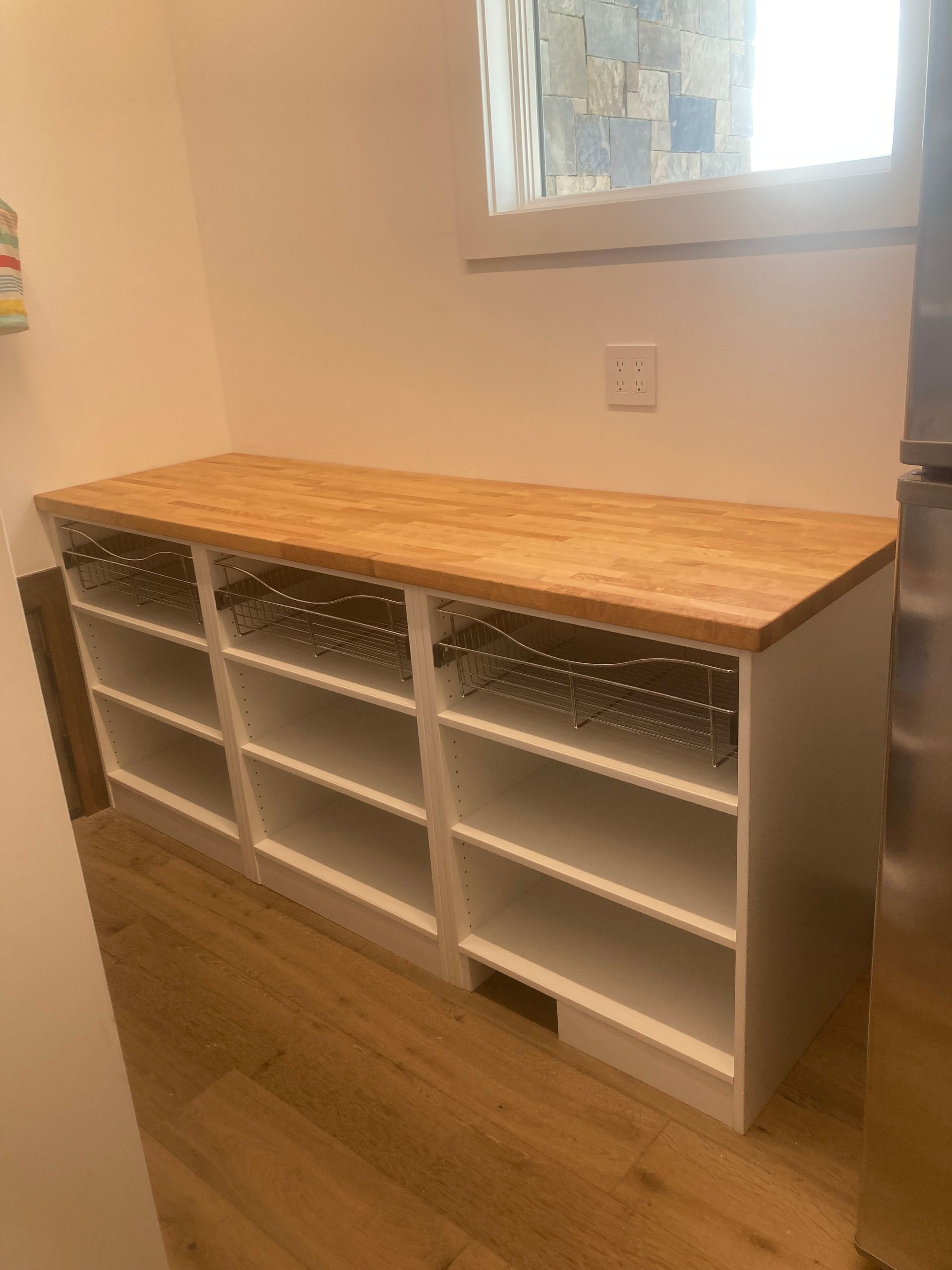 White storage bench with wooden countertop. Shelves below with wire baskets. Located near a window.