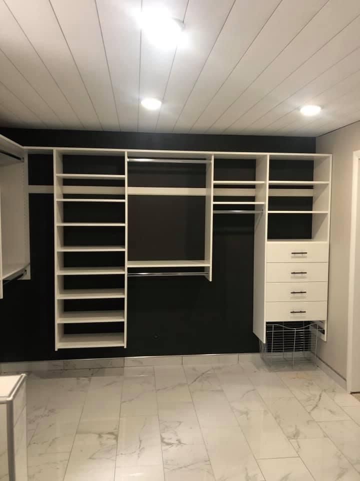 White closet organizer against a black wall, shelves, drawers, and hanging rods, in a room with marble flooring.