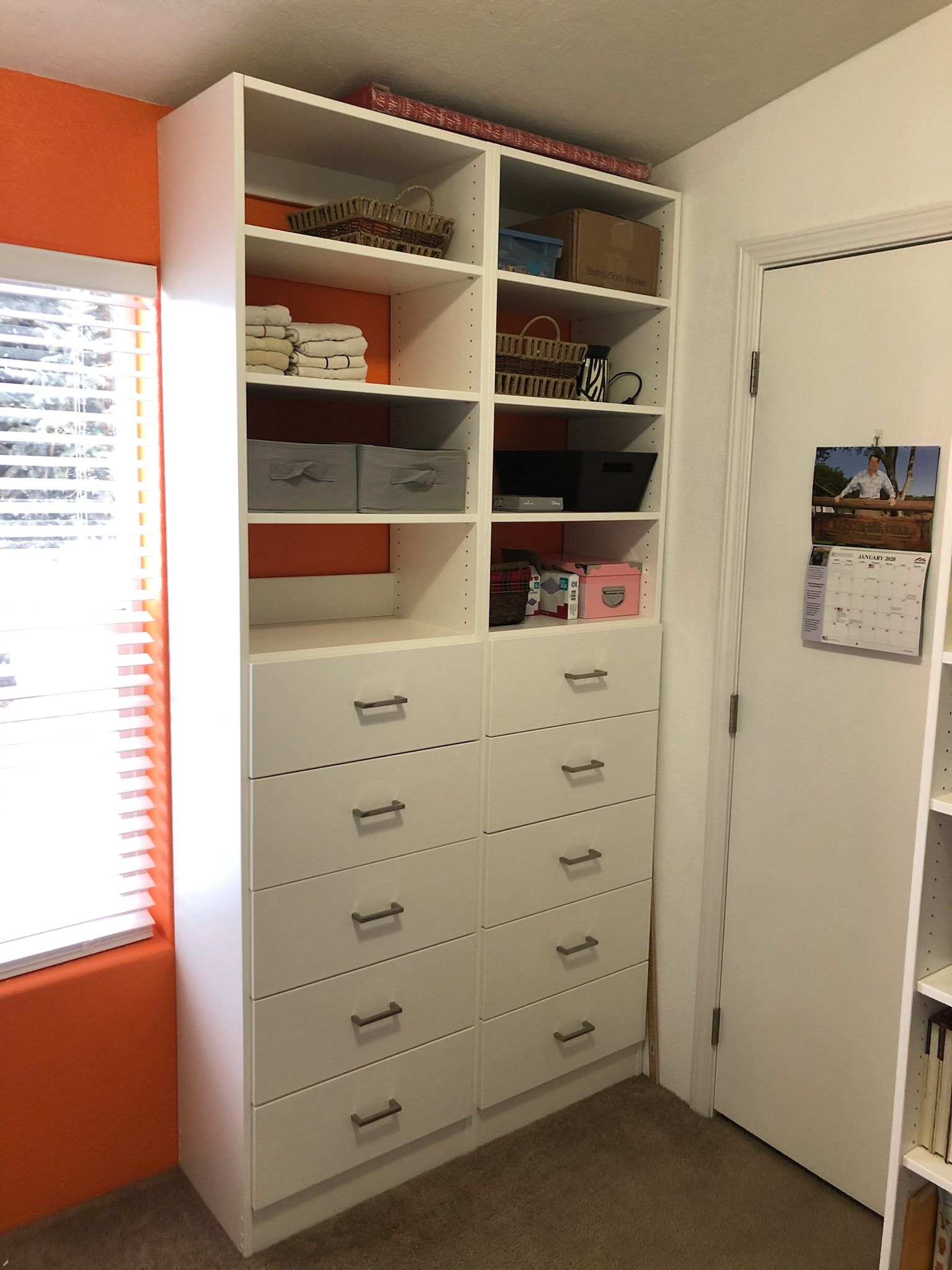 White shelving unit with drawers and shelves, next to an orange wall and a white door.