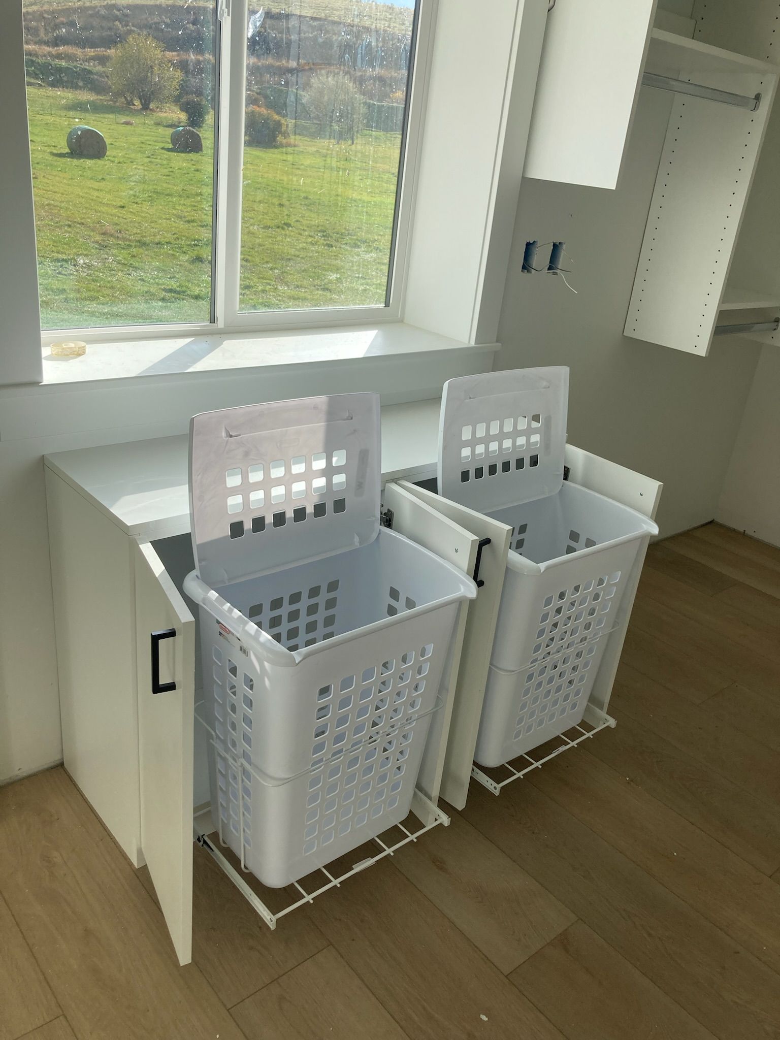 White laundry hamper pull-out baskets in a cabinet under a window, in a light-colored room.