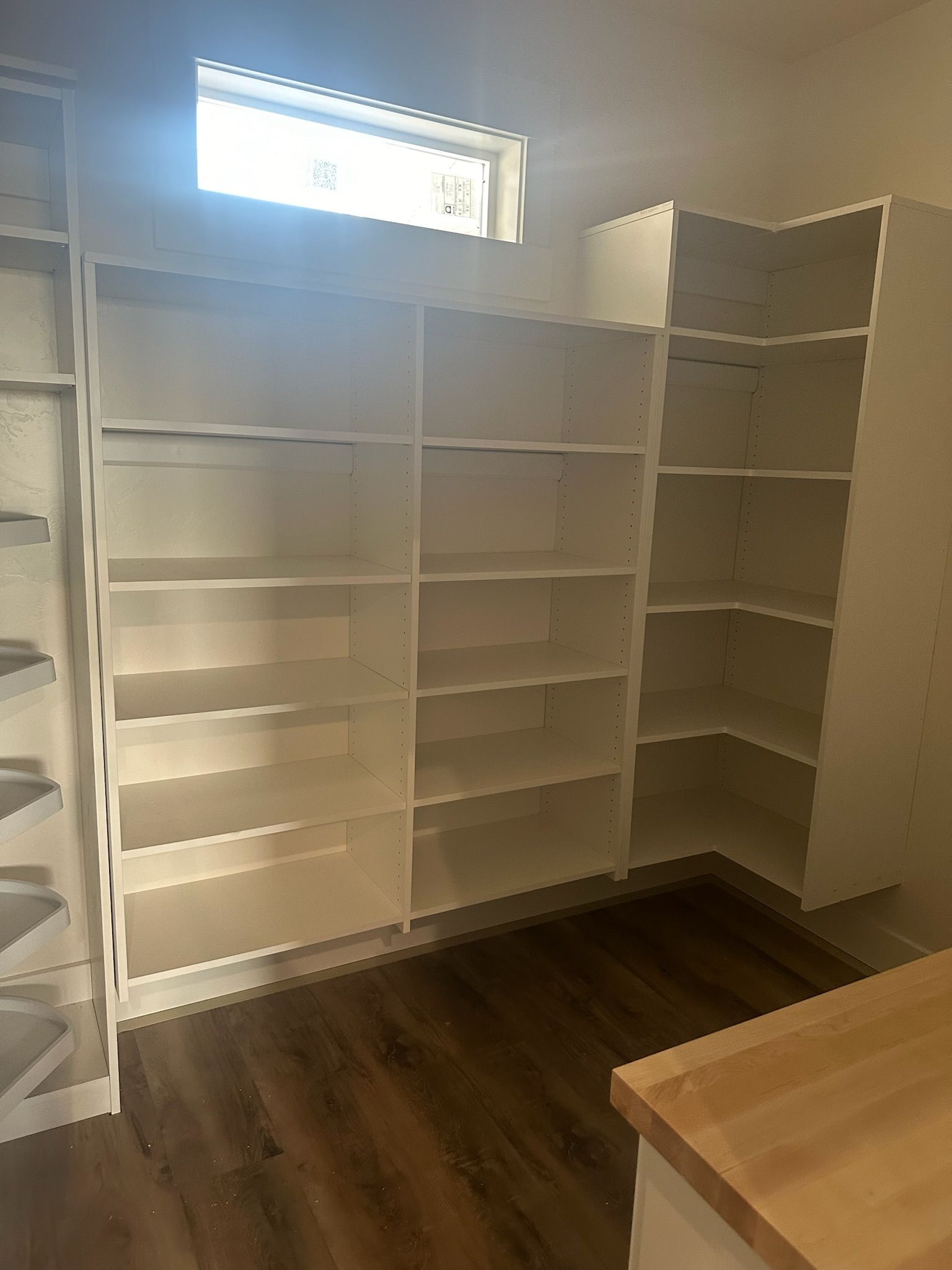 White built-in shelves in a pantry with a corner design, under a small window. Wood-look flooring.