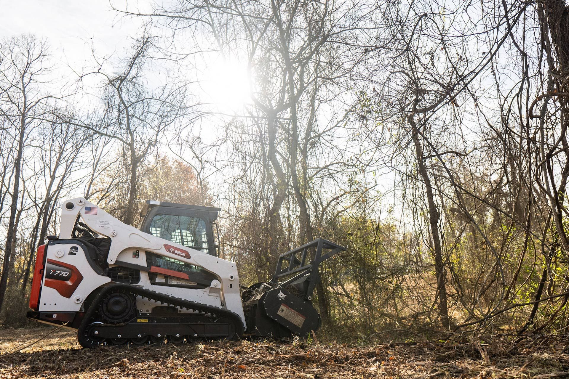 Bobcat clearing brush in a wooded area; sunny.