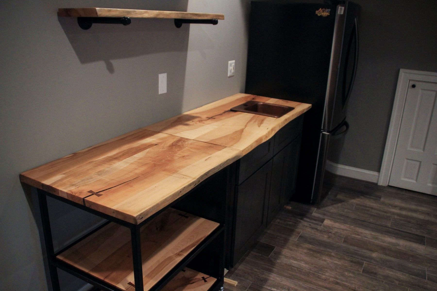 Kitchenette with wooden counter, black cabinets, and a refrigerator. Shelf above.