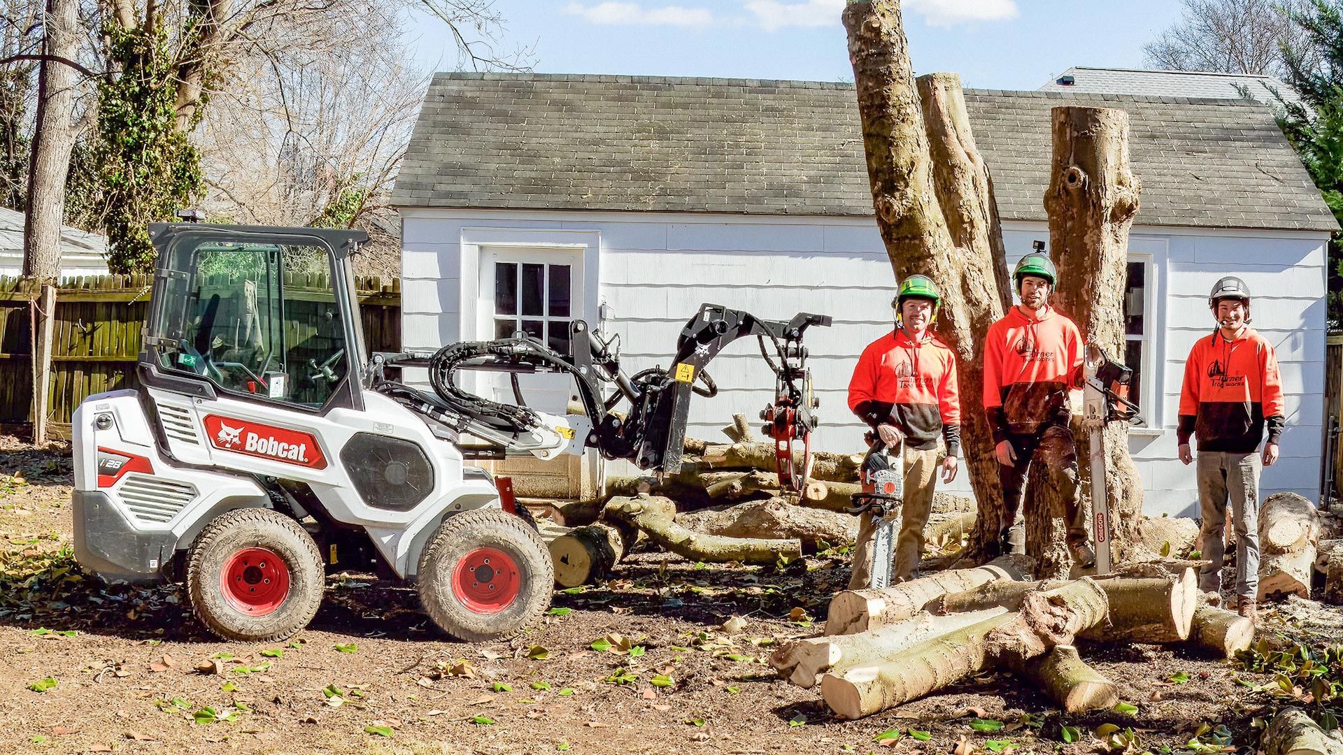 Three tree service workers stand next to a white Bobcat in front of a house, with logs and cut tree trunks.