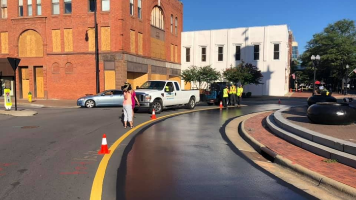 Construction workers pave a section of a curved city street near red brick and white buildings on a sunny day.