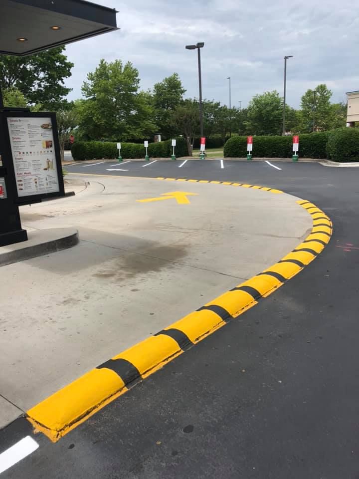 A yellow and black speed bump curving along the pavement at a drive-thru, with a menu board and parking spots in the back.
