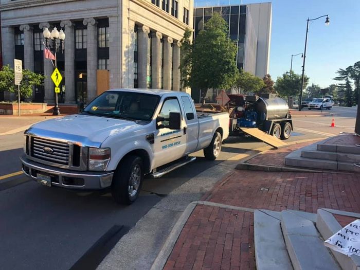 A white pickup truck pulling a trailer with a large tank parked on a city street next to a brick building.
