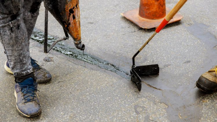 A worker uses a specialized nozzle and a handheld tool to apply hot sealant into a crack in a paved road.
