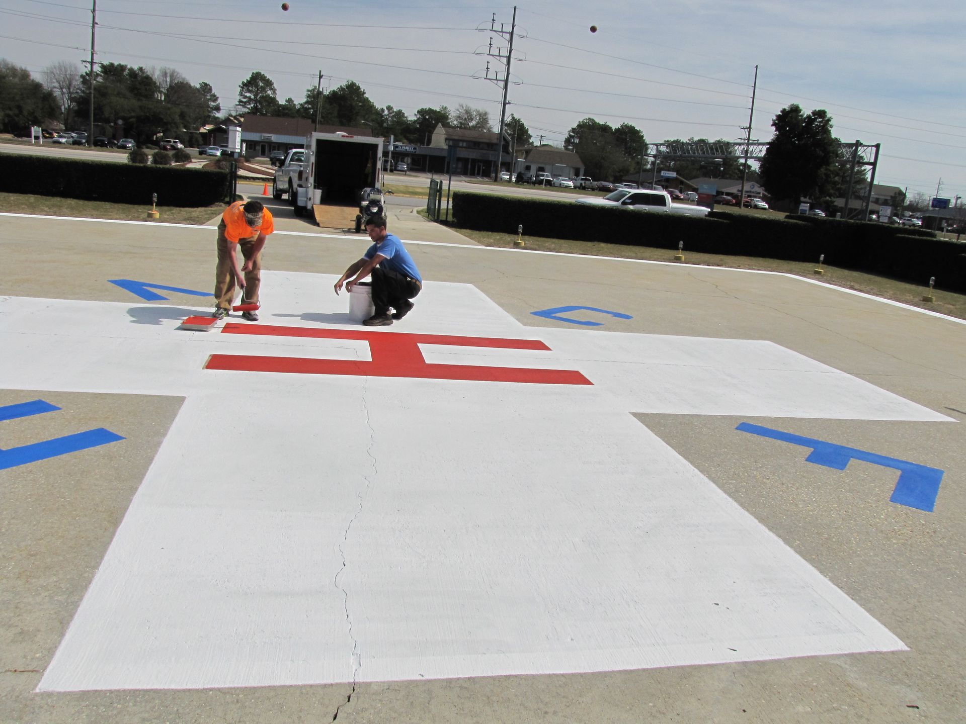Two workers painting a large white surface with red and blue markings outdoors.