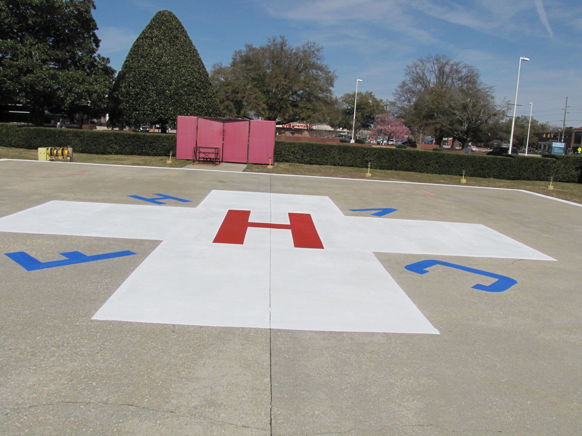 Helipad marking with red H on white pavement, blue directional arrows, and pink barricades in a park