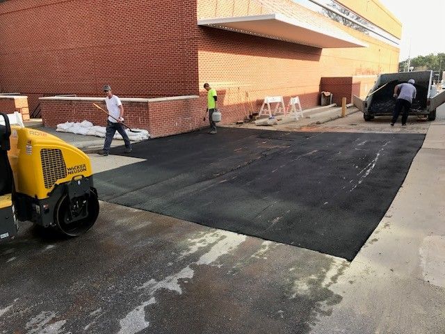 Workers paving a black asphalt driveway beside a brick building with a yellow roller in the foreground