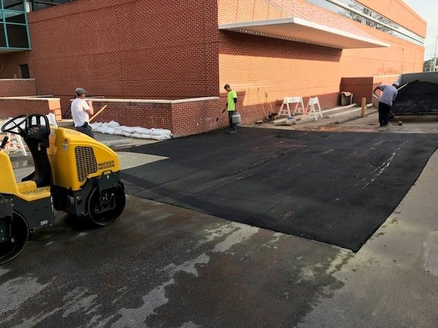Workers paving a black asphalt ramp beside a brick building, with a compact roller and cones nearby