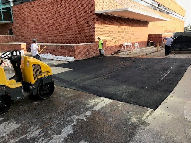 Construction workers paving a black asphalt walkway beside a brick building with a yellow roller in front