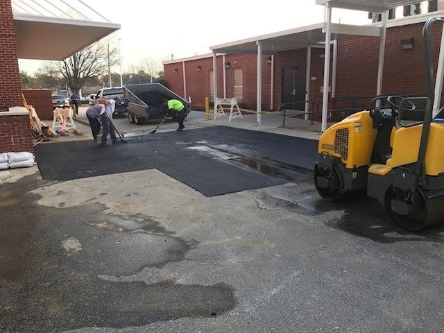 Workers paving asphalt in a parking lot beside a brick building, with a yellow roller nearby