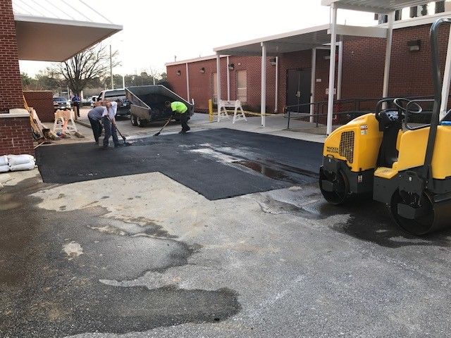 Road crew paving a school courtyard beside a yellow roller and brick buildings
