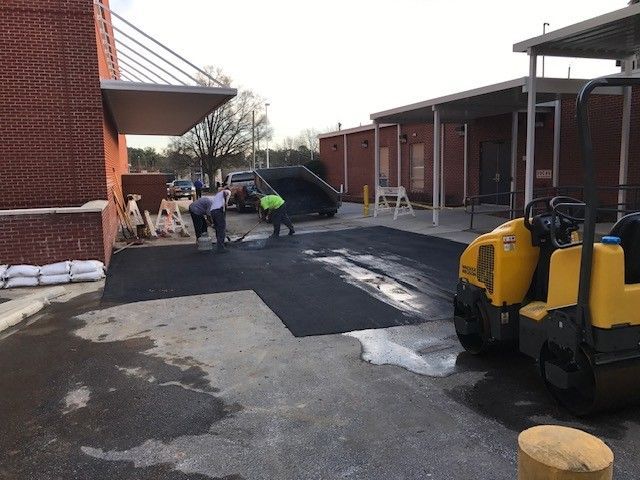 Workers paving asphalt beside a brick building, with a yellow machine in the foreground