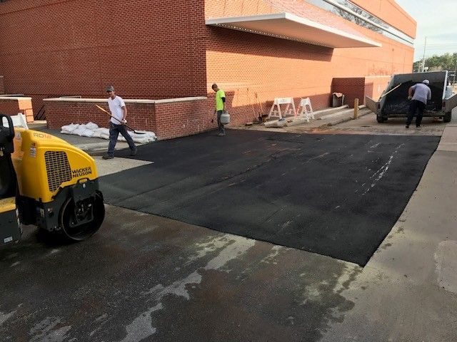 Workers paving a black asphalt patch beside a red brick building, with a yellow machine in the foreground.