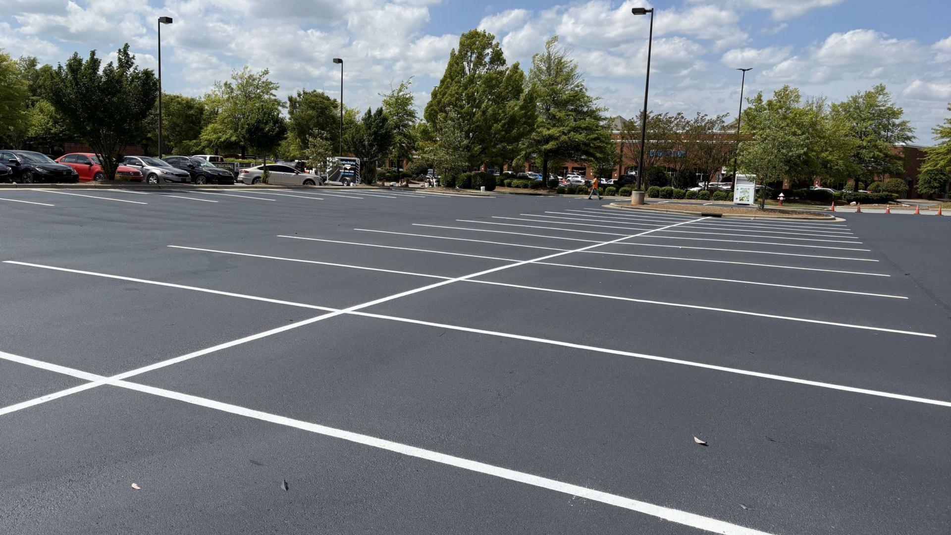 A freshly paved parking lot with white painted stalls, trees, and light poles under a partly cloudy sky.