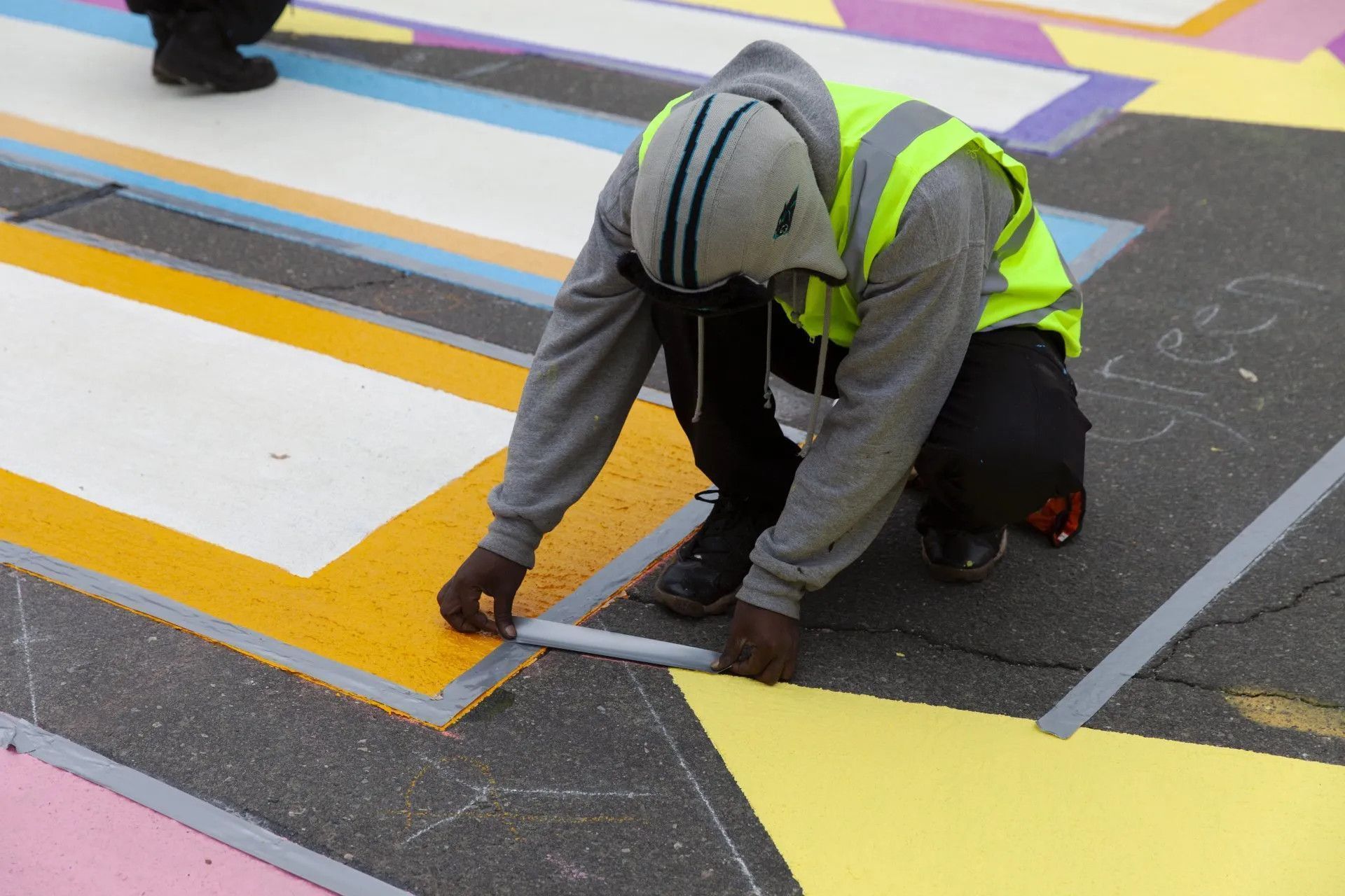 A person wearing a high-visibility vest uses painter's tape to outline a colorful, geometric street mural.