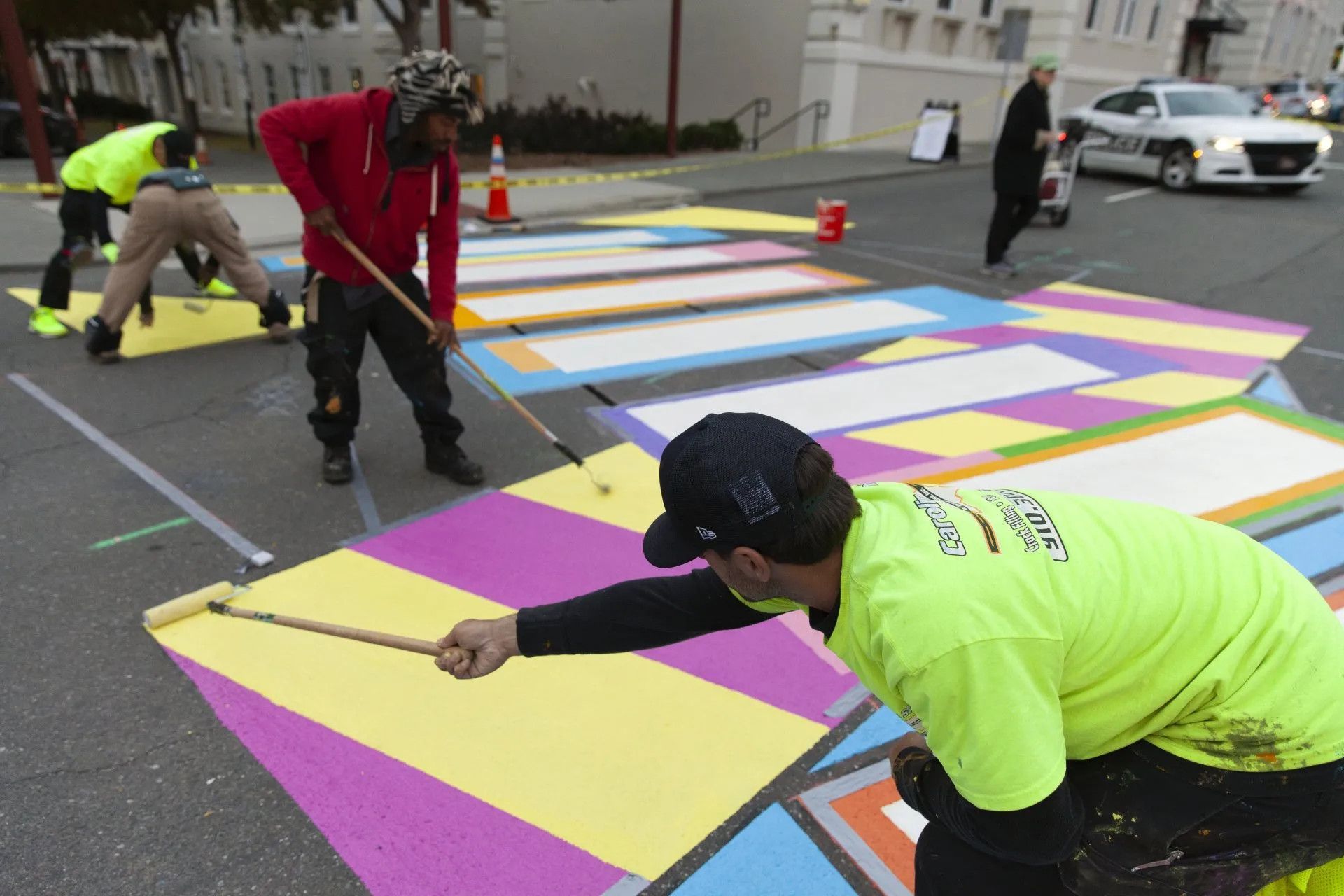 People in bright shirts paint a colorful, geometric mural onto an asphalt street crosswalk while a police car is nearby.