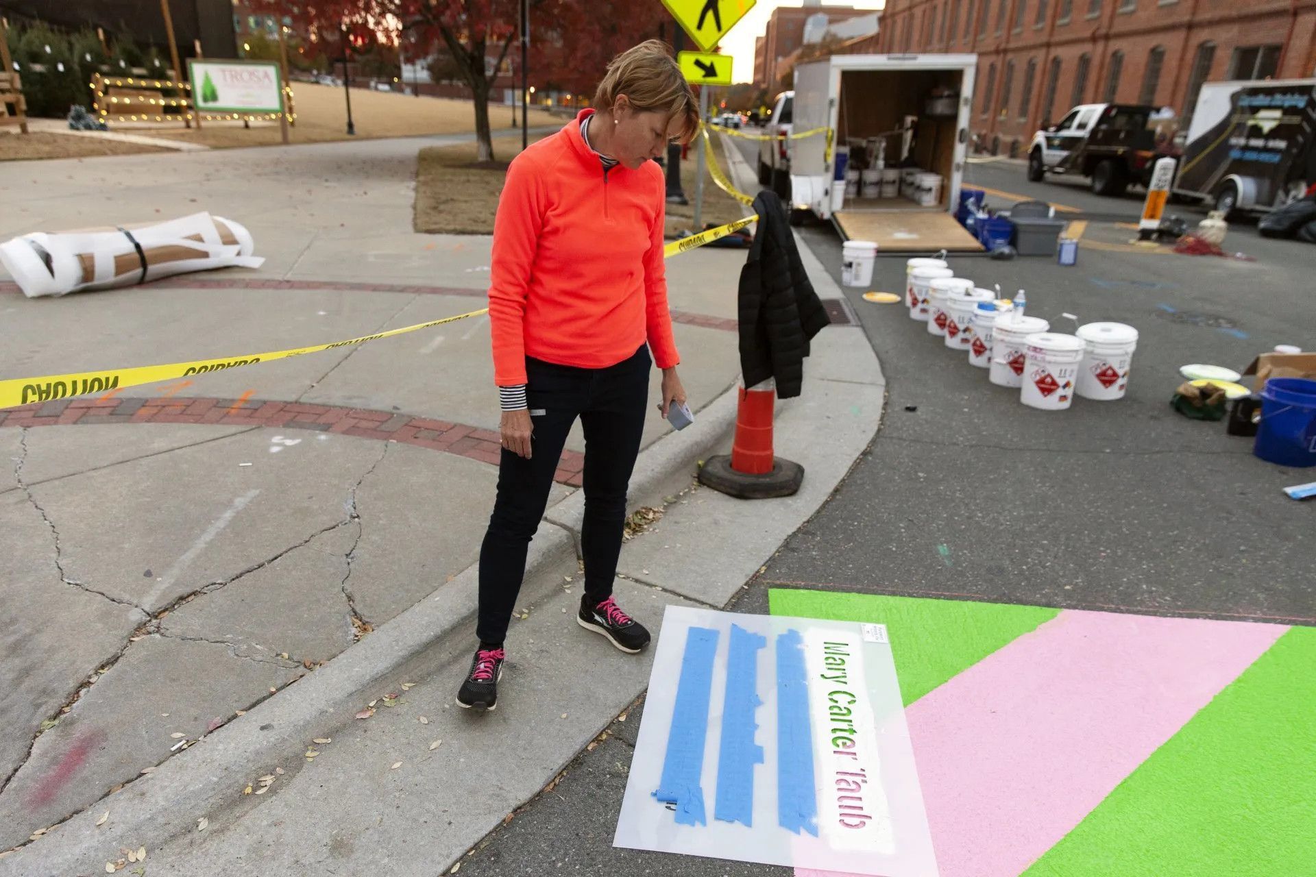 A person in a bright orange shirt and black pants stands in a parking lot next to painted pavement and rows of paint buckets.