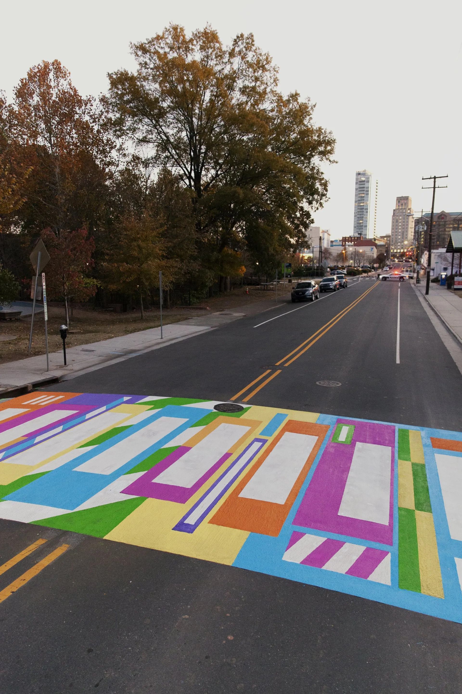 A crosswalk painted with a colorful, geometric pattern on an asphalt road near trees and city buildings.