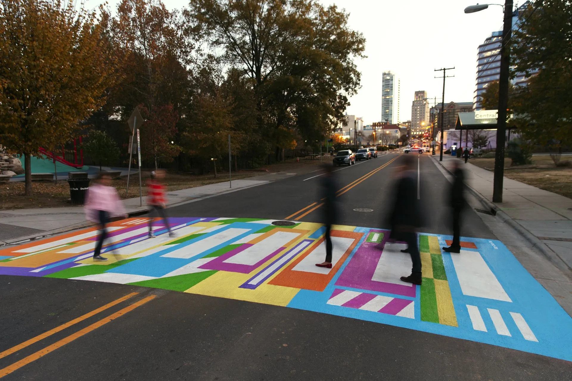 Blurred pedestrians cross a city street marked by a vibrant, multi-colored geometric mural crosswalk at dusk.