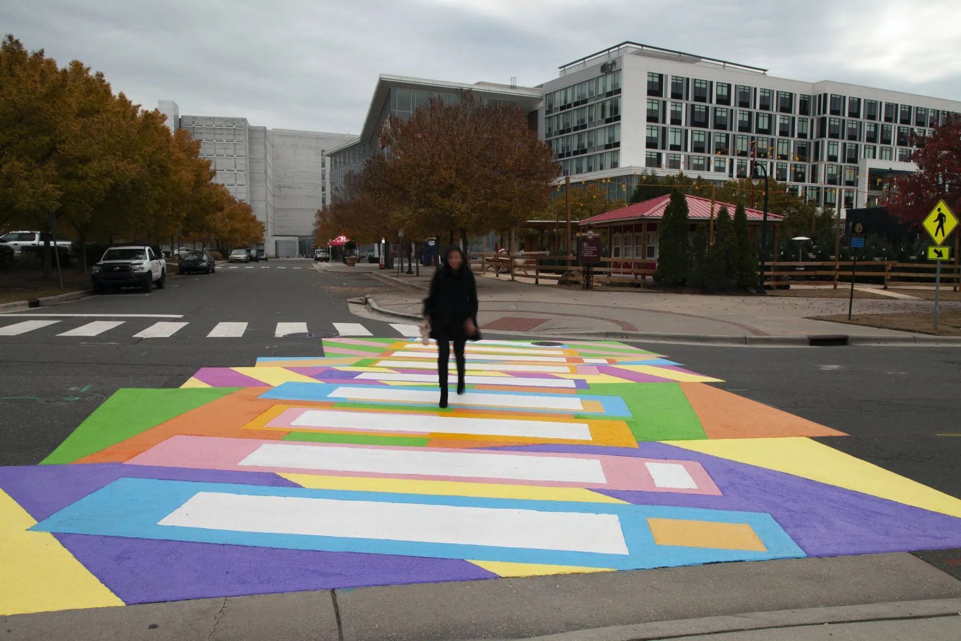 A person walks across a vibrant, multicolored painted crosswalk on an asphalt street near modern buildings and trees.