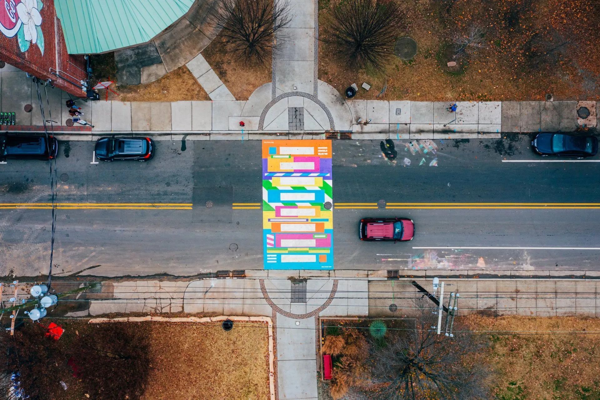 An aerial view of a multi-colored, striped crosswalk on a gray asphalt street between two sidewalks.