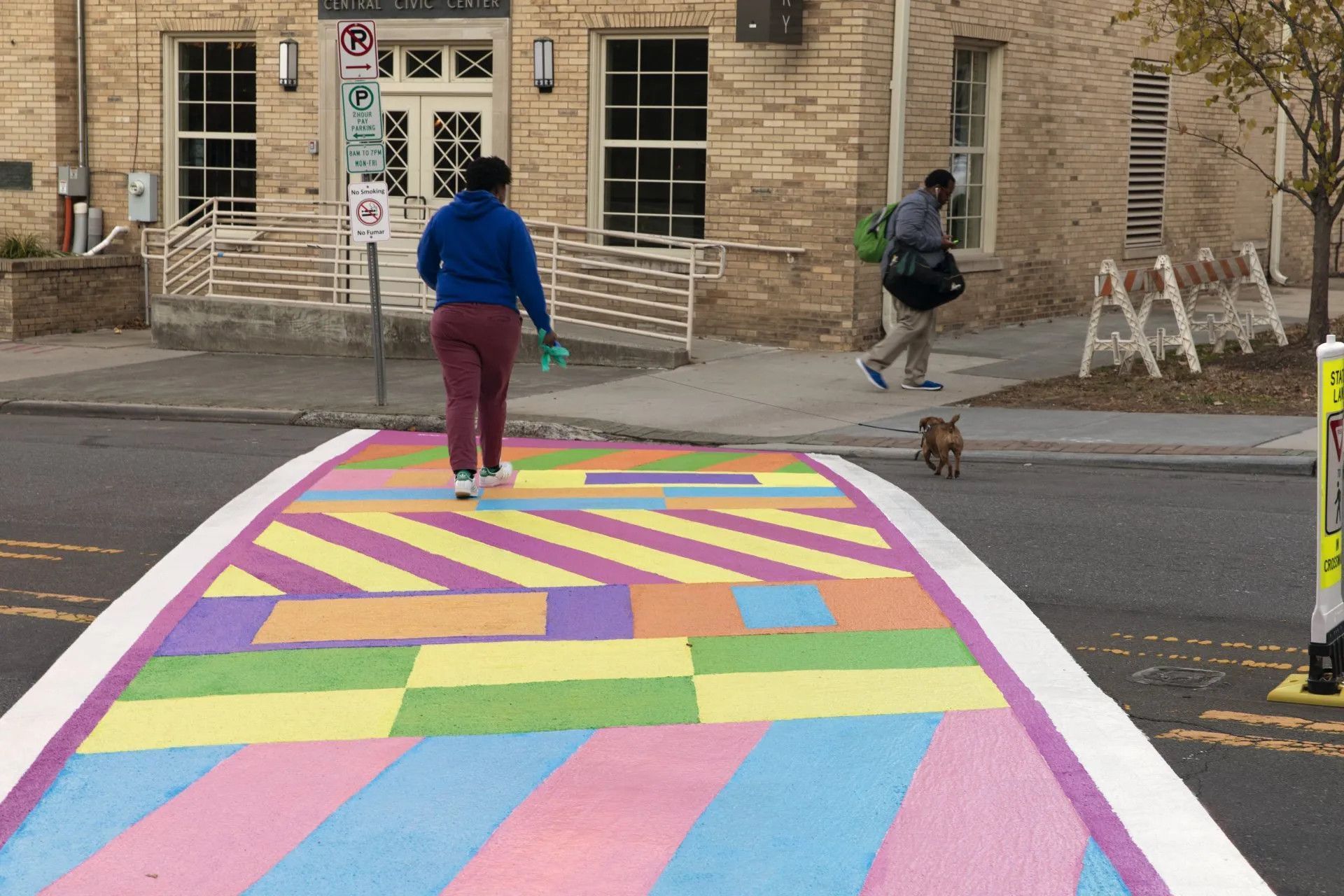 A person in a blue hoodie walks across a colorful painted crosswalk, with a small dog walking alongside on the pavement.