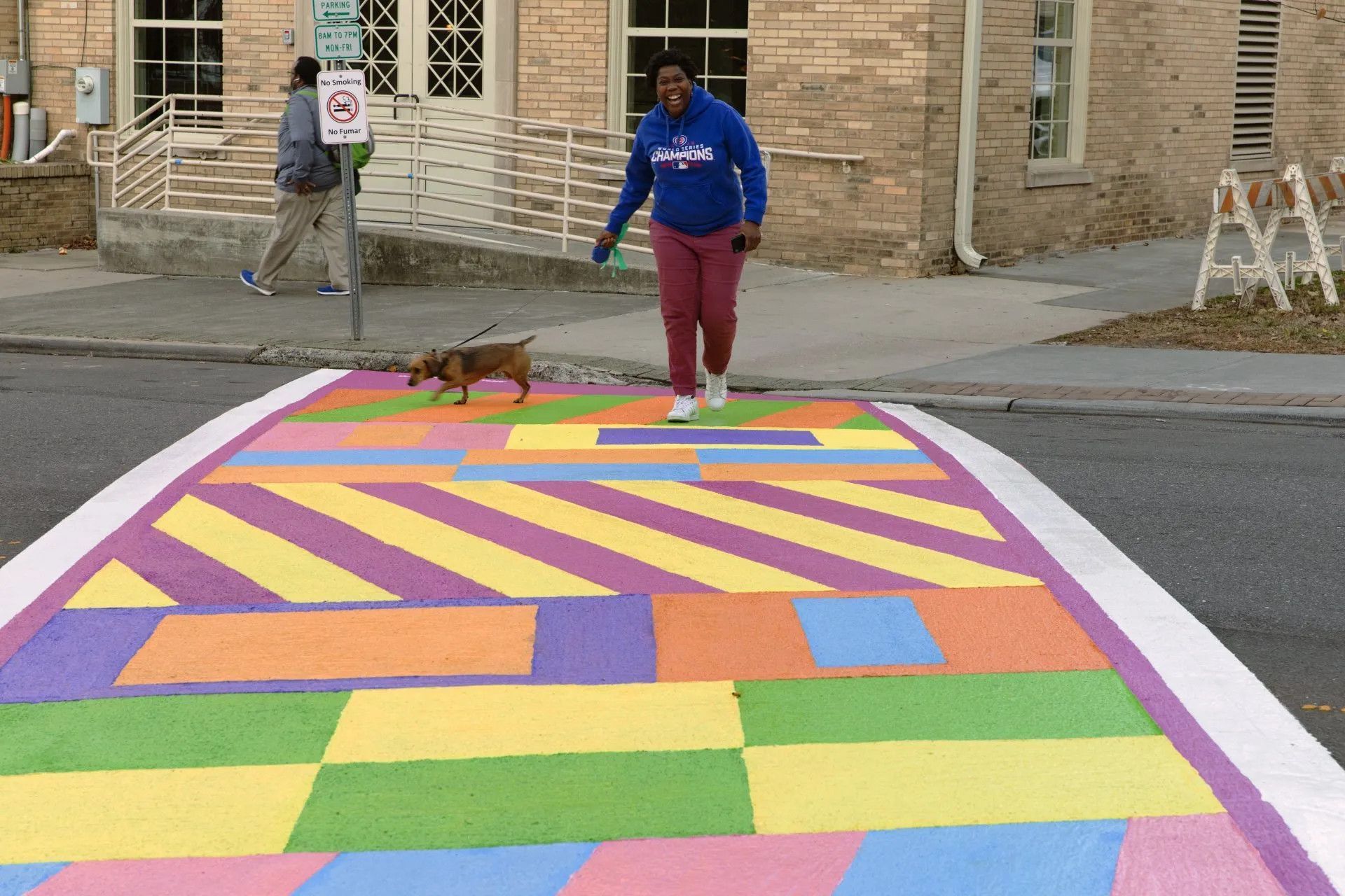 A person walks a small brown dog across a colorful, geometric-patterned crosswalk in front of a brick building.