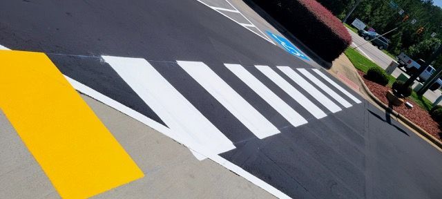 Painted crosswalk with white stripes and a yellow curb on a slanted street corner.