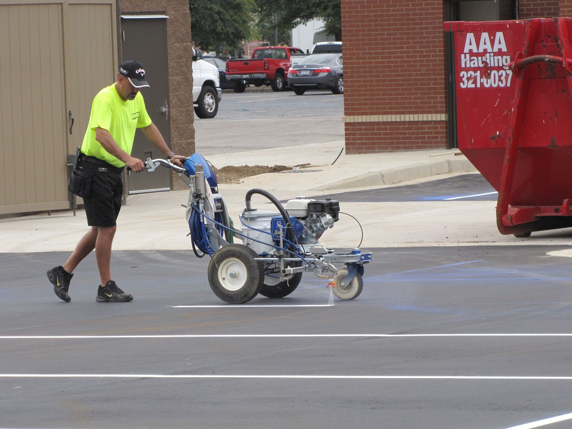 Worker in neon shirt pushing a blue line-marking machine on a street near a red dumpster.