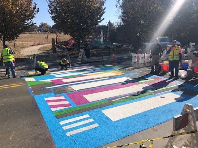 People painting a large colorful street mural on a road, with volunteers and supplies nearby.