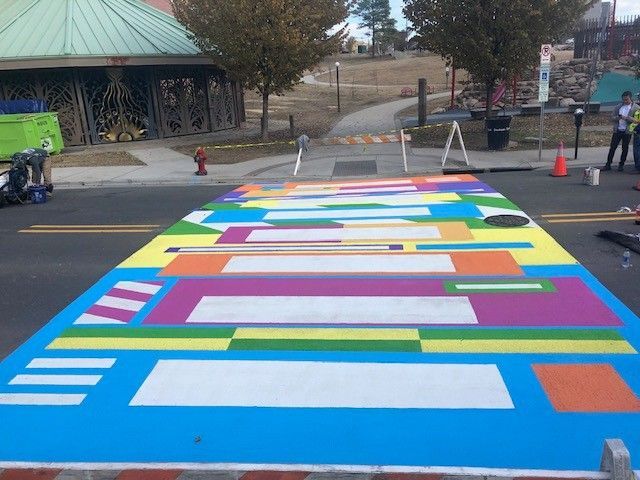 Colorful geometric street mural painted across a crosswalk in a parking lot near a pavilion