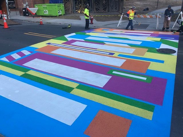 Colorful striped street mural being painted on a roadway, with workers and traffic cones nearby