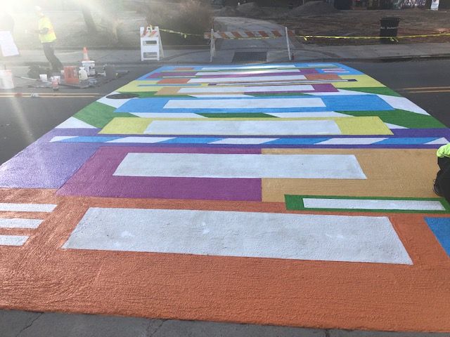 Colorful painted crosswalk mural on a road, with geometric shapes and street barriers in the background.