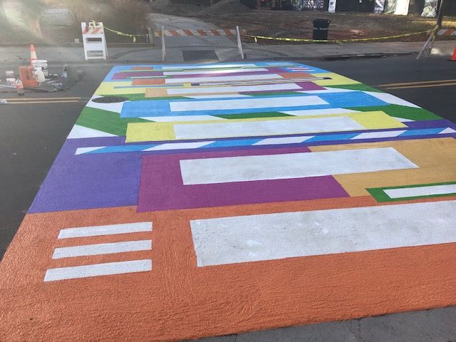 Colorful geometric street mural painted across a road crossing, with bold blue, purple, green, orange, and white shapes.