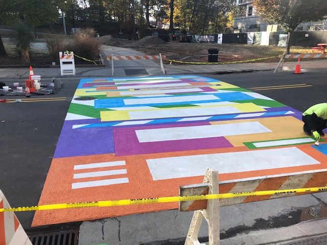 Colorful painted street mural on a closed road, with workers adding details and traffic cones nearby