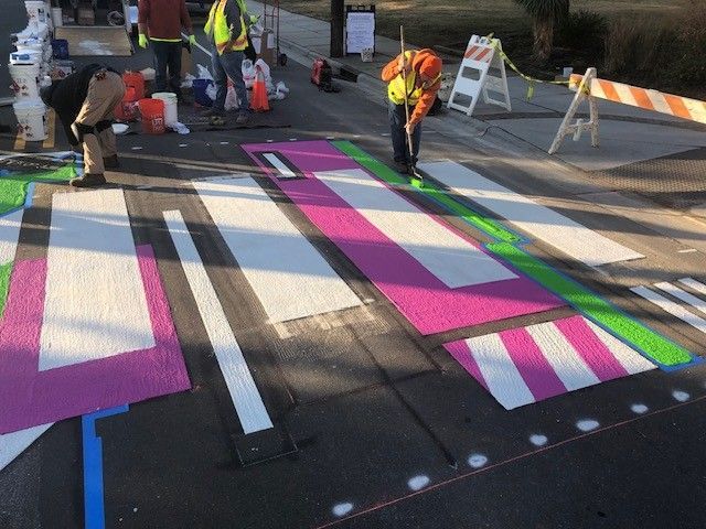 Workers painting bright pink and green road markings on a crosswalk during street maintenance